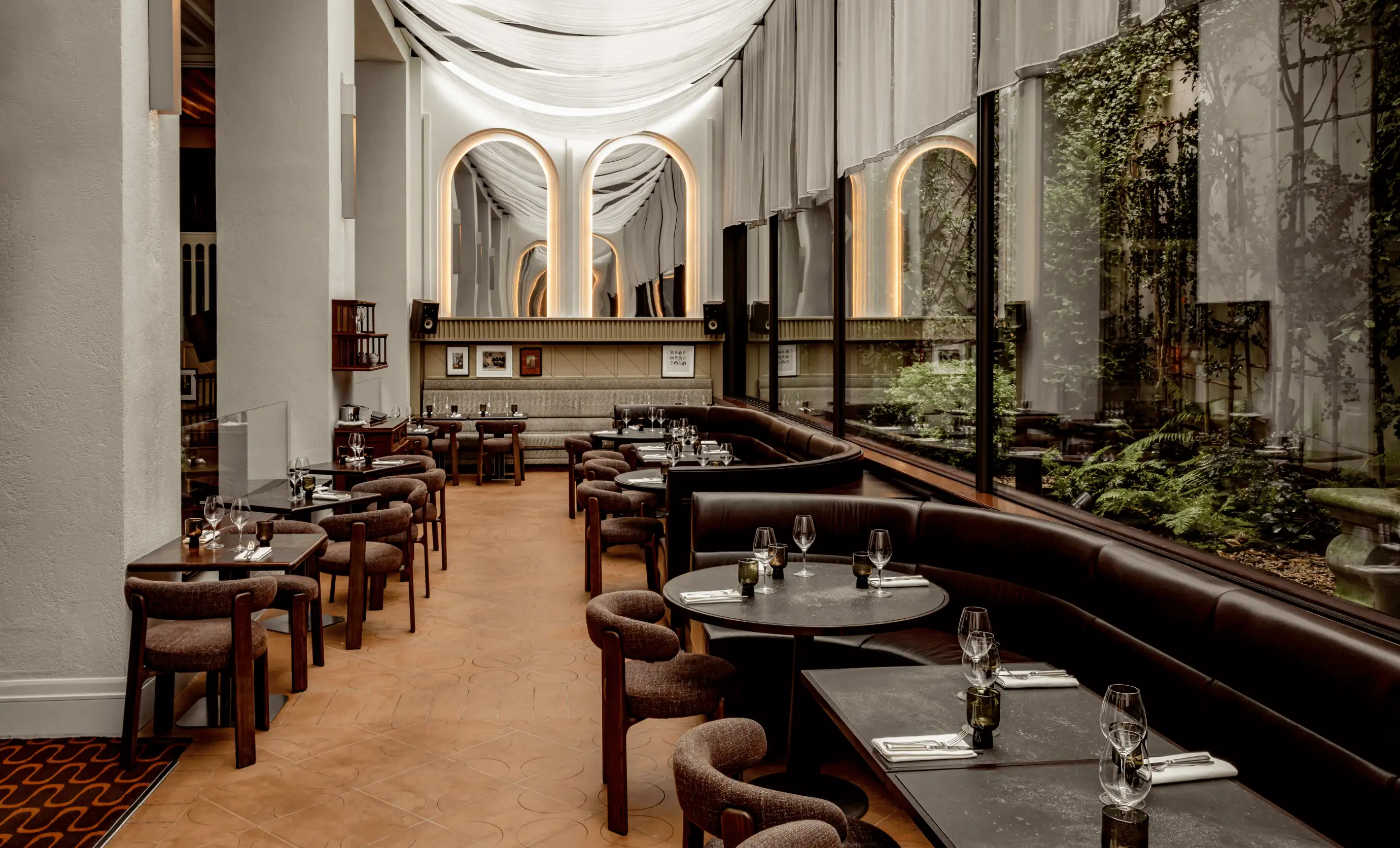 Modern indoor restaurant seating with round and square tables, brown chairs, leather banquette, and large windows overlooking a green courtyard.