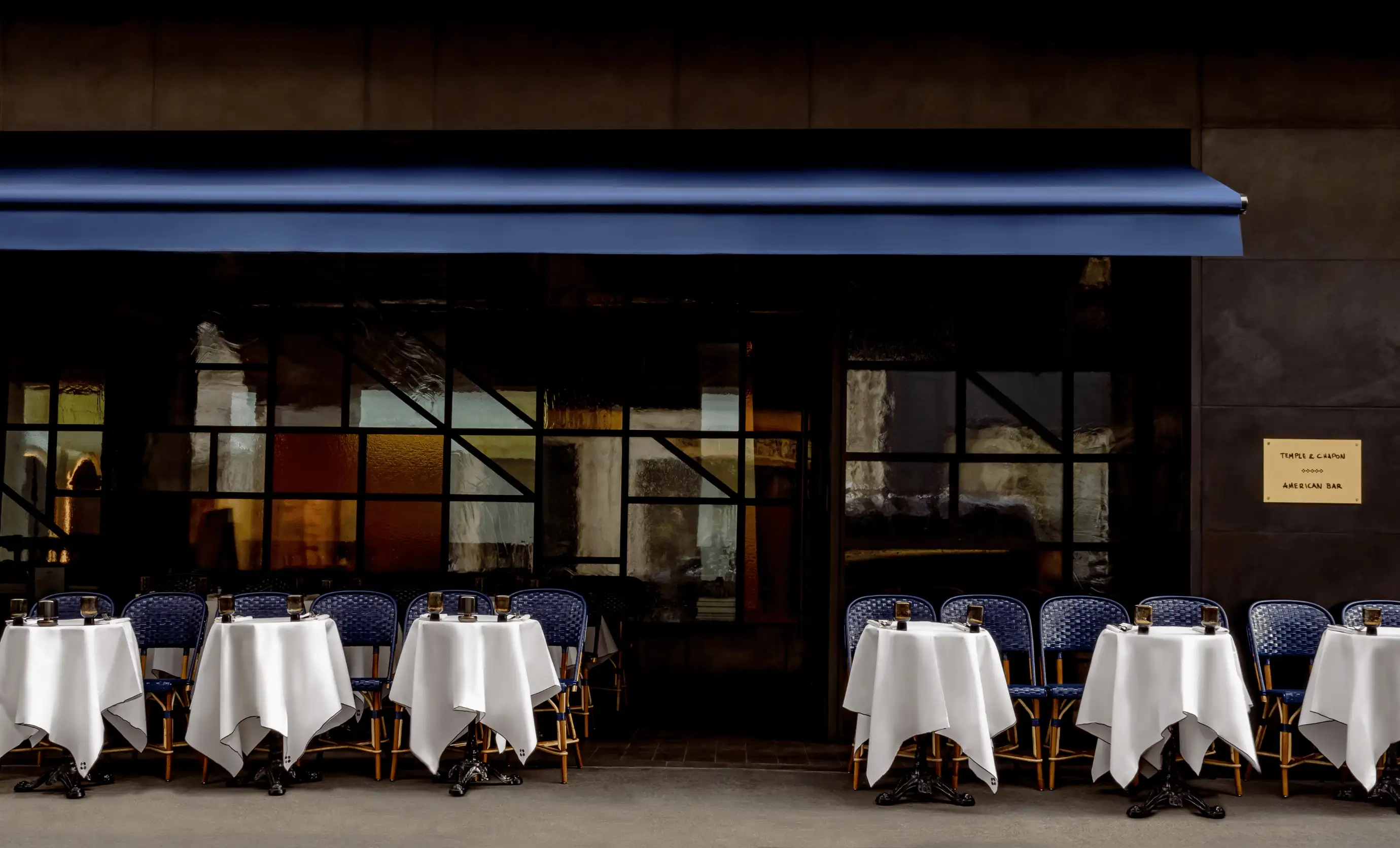 Outside seating area of a restaurant with small tables covered in white tablecloths and blue chairs under a blue awning.