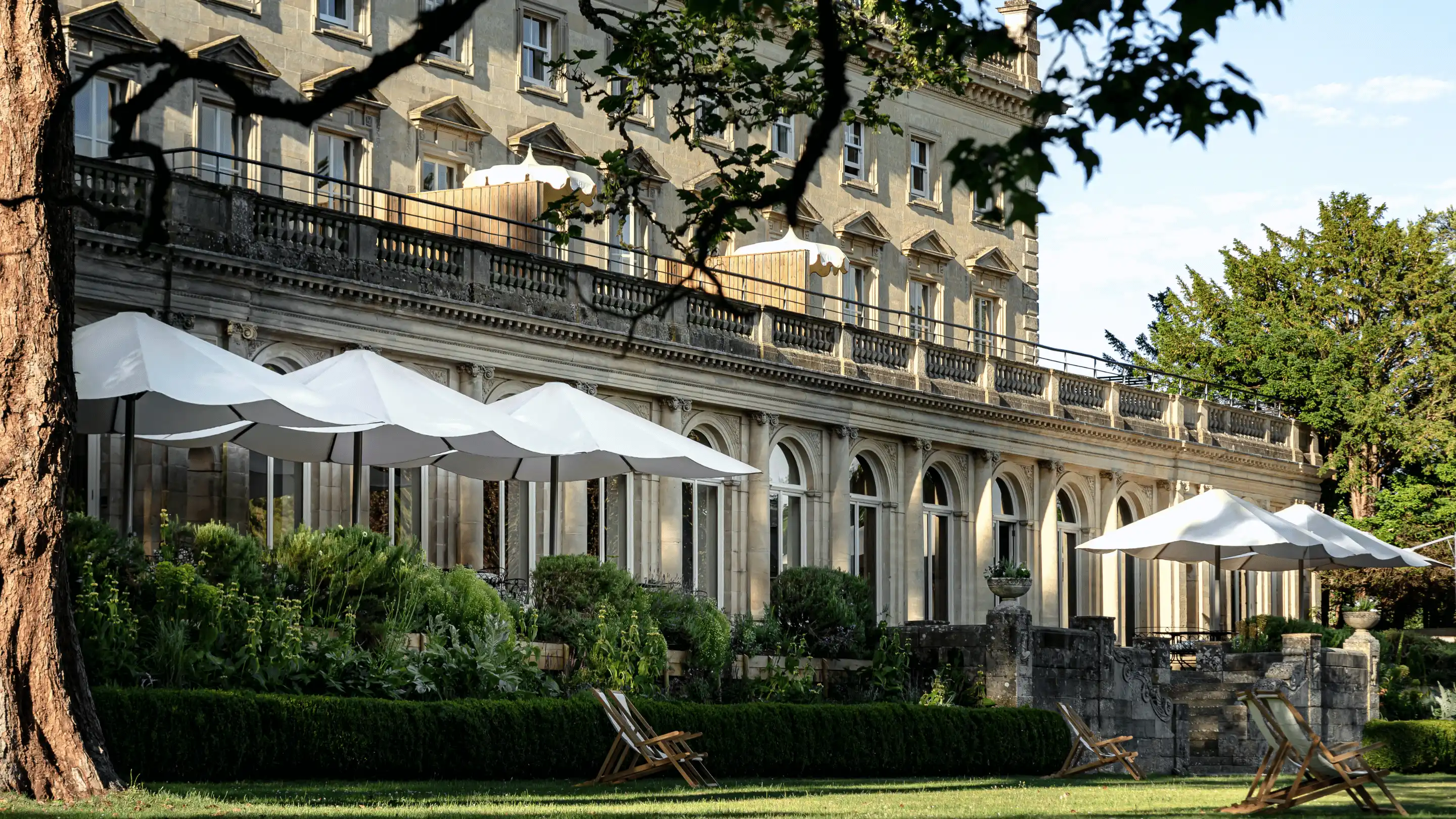 Historic stone hotel building with arched windows, white umbrellas, garden plants, and wooden deck chairs on the lawn.