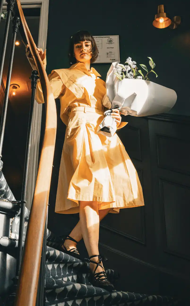 Mujer con un vestido amarillo bajando escaleras alfombradas con un ramo de flores blancas envueltas en papel.