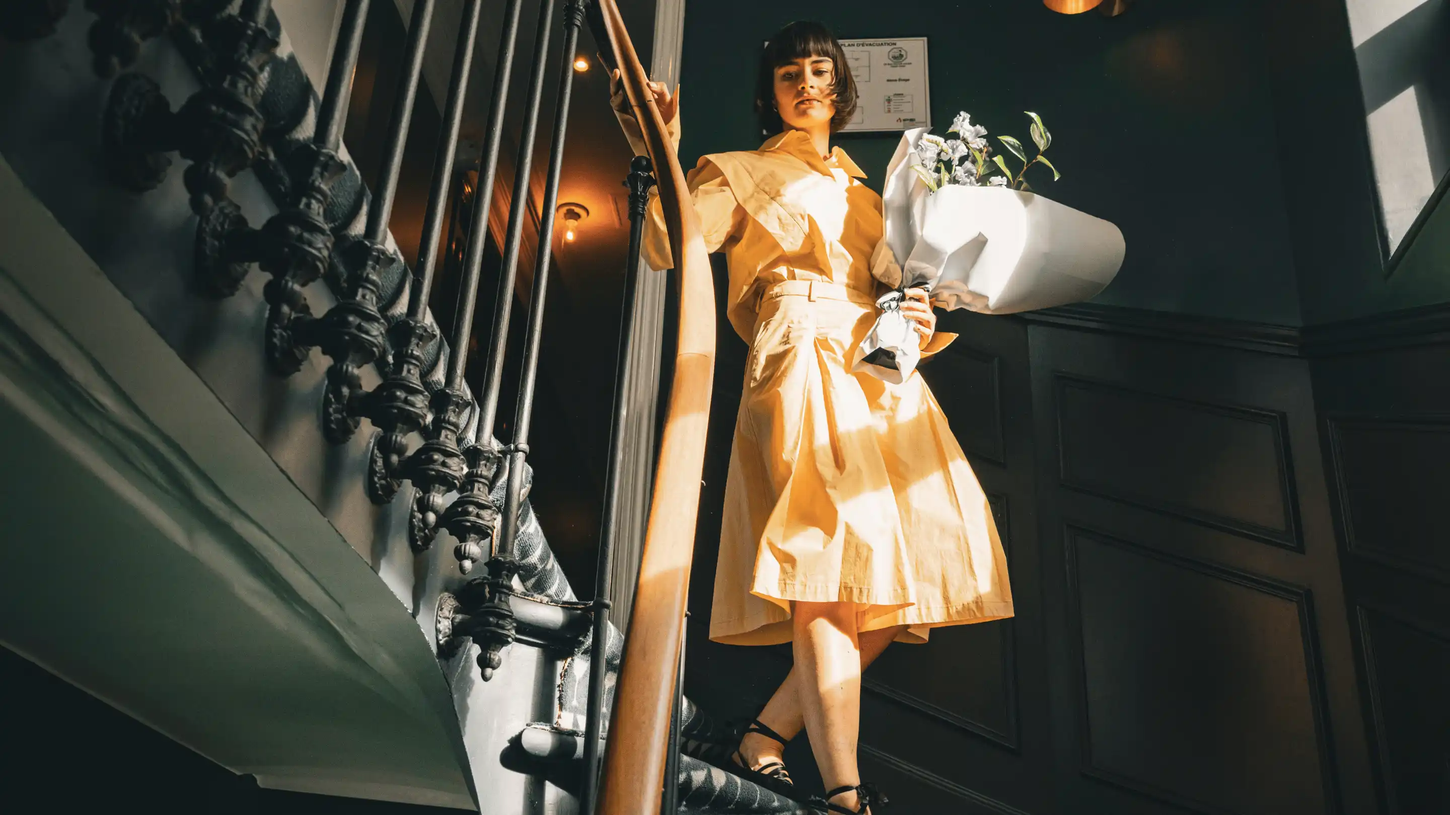 Mujer con un vestido naranja claro que sostiene un ramo de flores blancas mientras está de pie en una escalera con paneles de madera oscura.