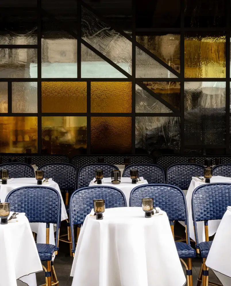 Table seating in a restaurant with white tablecloths, dark wicker chairs, and a decorative glass paneled wall with amber and clear sections.