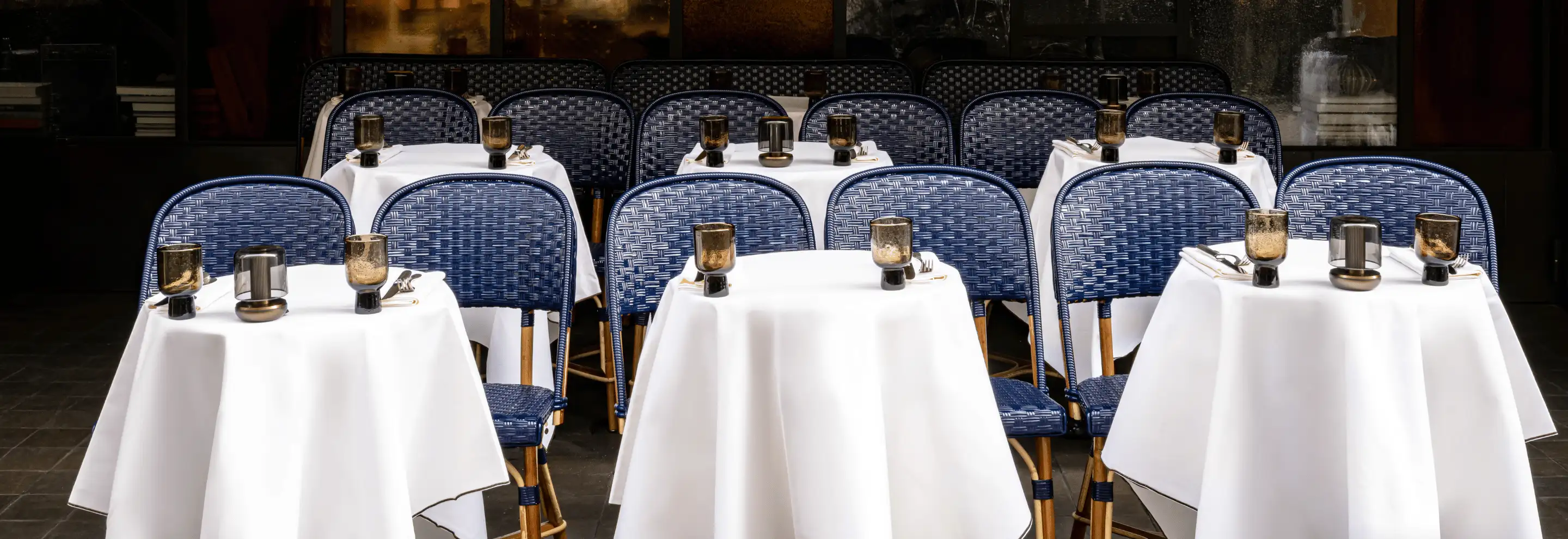 Neatly arranged outdoor restaurant tables with white tablecloths, blue woven chairs, and gold-rimmed glassware.