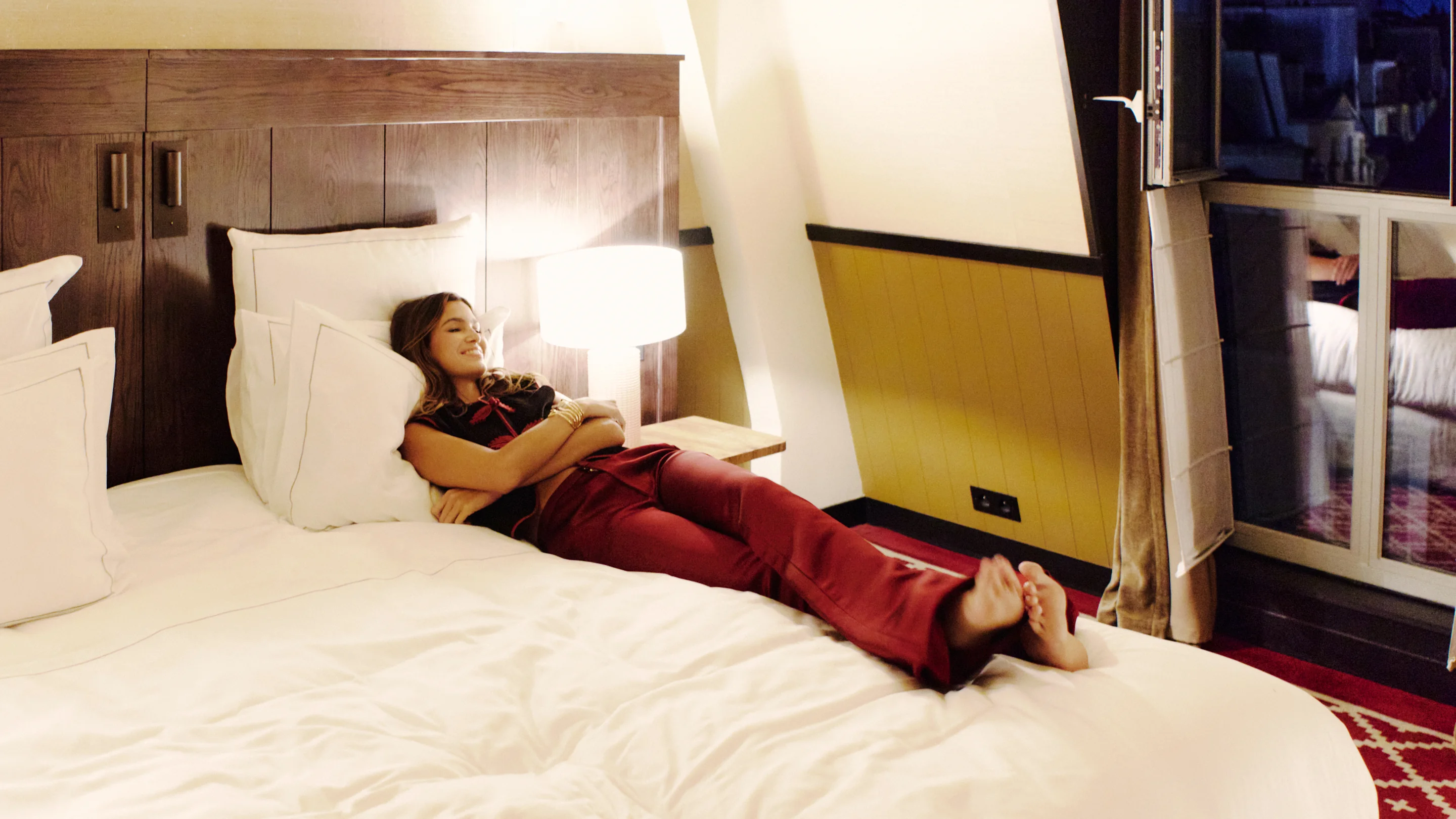 Woman in red pants reclining on a white bed in a warmly lit hotel room with wooden headboard and bedside lamp.