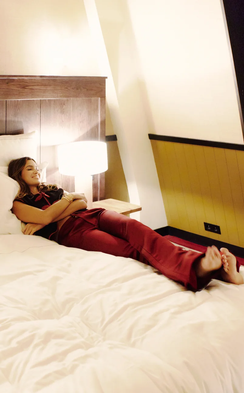 Woman reclining comfortably on a bed with white bedding in a warmly lit hotel room.