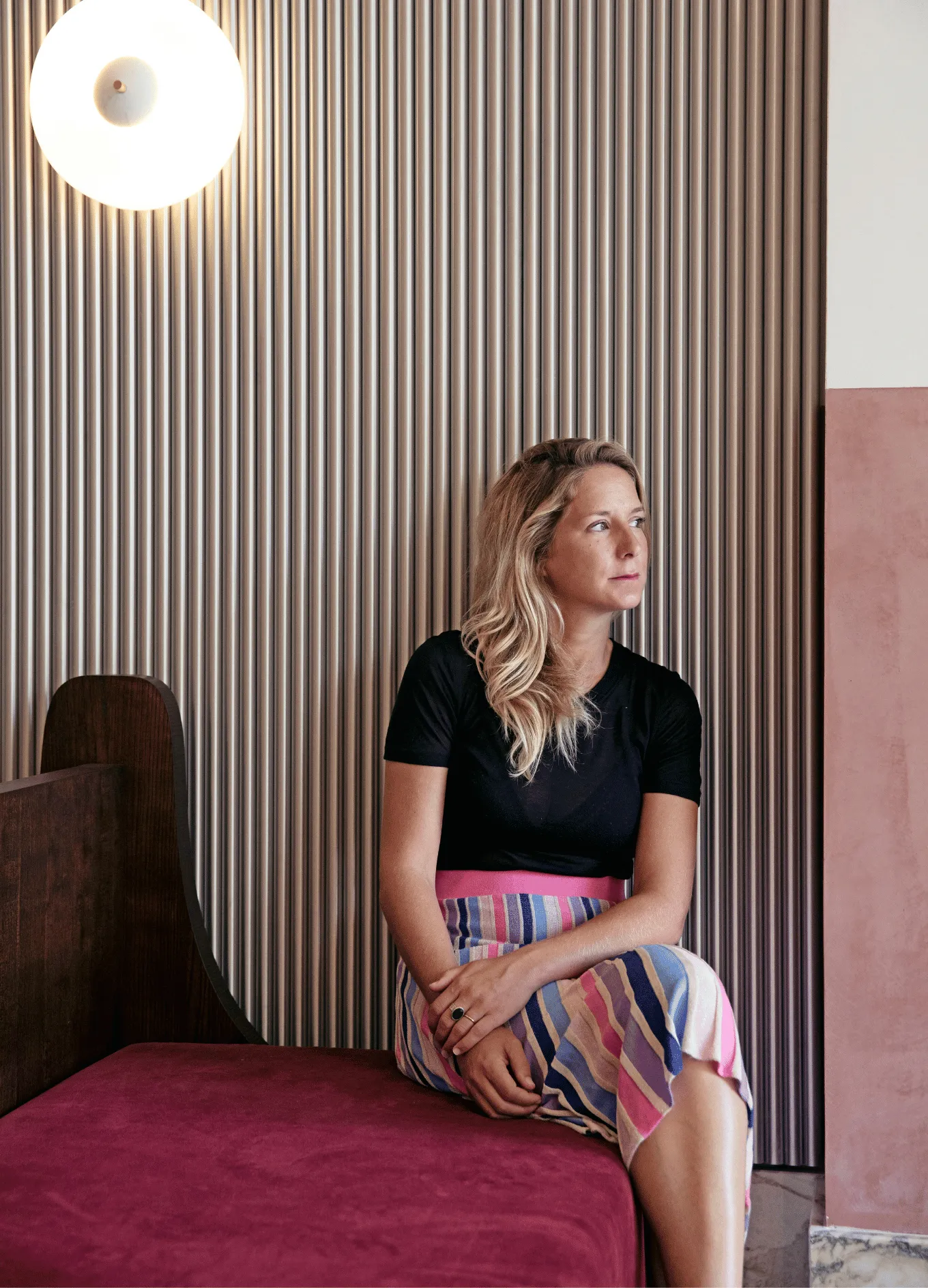 Woman with blonde hair in a black top and colorful striped skirt sitting on a maroon bench against a striped wall with a round light fixture.