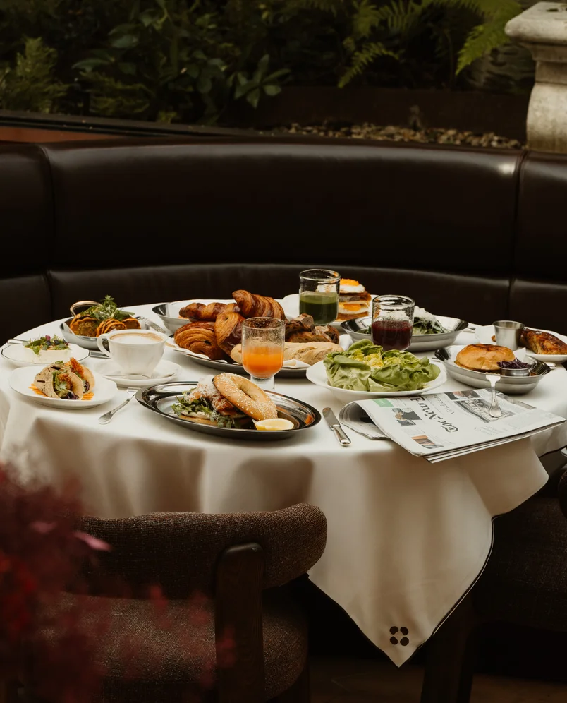 Round table with a white tablecloth set with various brunch dishes including croissants, sandwiches, salads, drinks, and a newspaper.