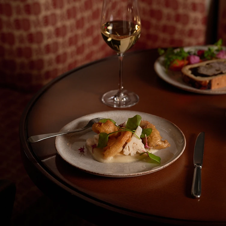 Plate with fried cauliflower florets on a creamy sauce, garnished with greens, beside a glass of white wine on a wooden table.