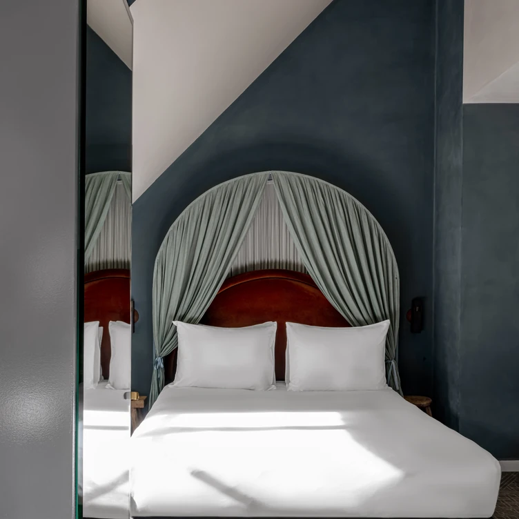 Modern hotel room with a double bed featuring white bedding and pillows, a curved dark-headboard framed by grey curtains, and dark blue walls.