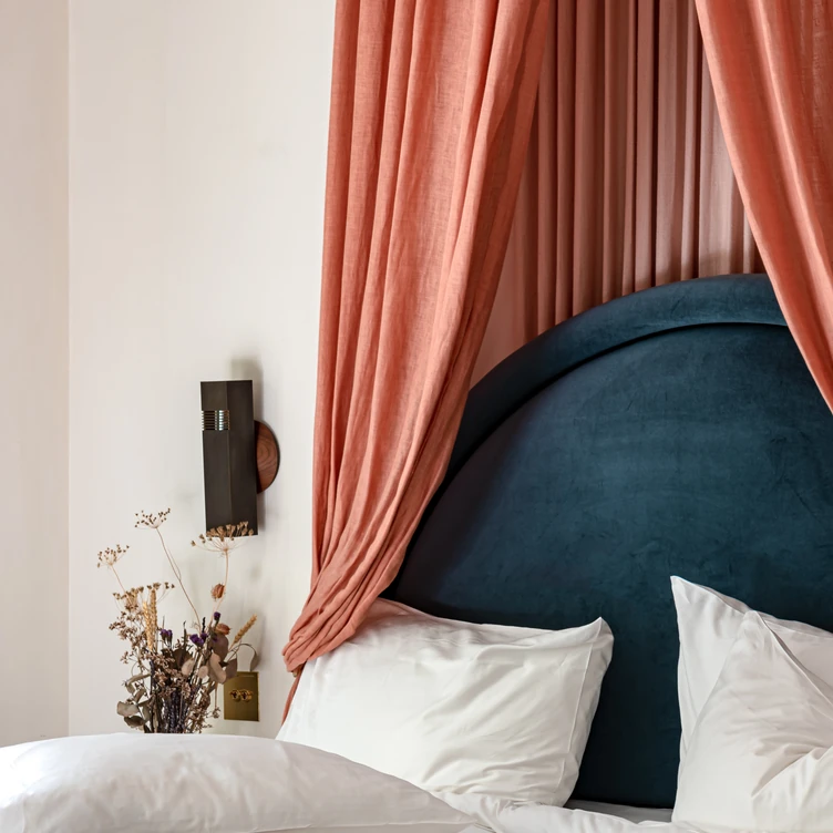 Close-up of a bed with white pillows, a teal rounded headboard, and peach curtains draped on either side.