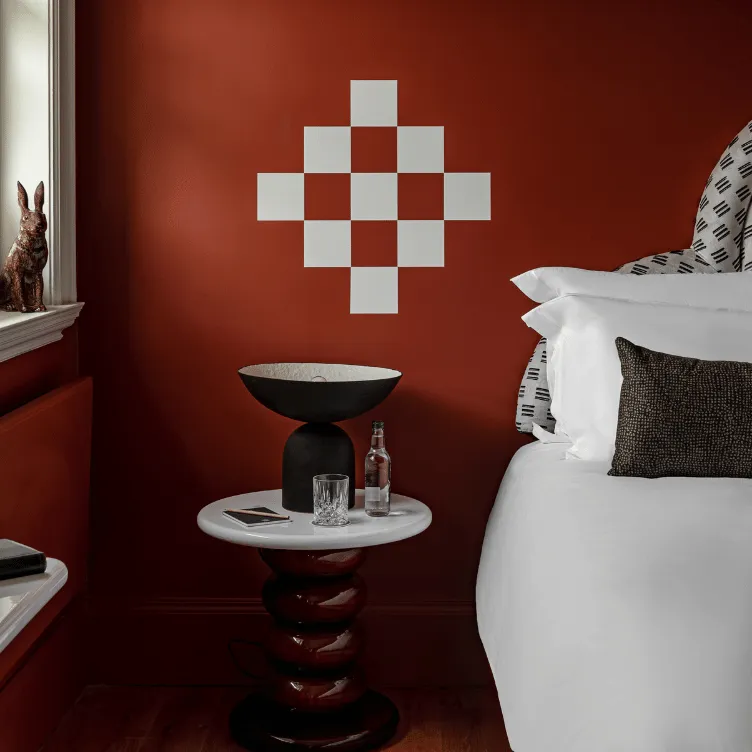 A bedside table with a black decorative bowl, a glass, and a bottle next to a bed with white linens, against a rust-colored wall with a white geometric wall decal.