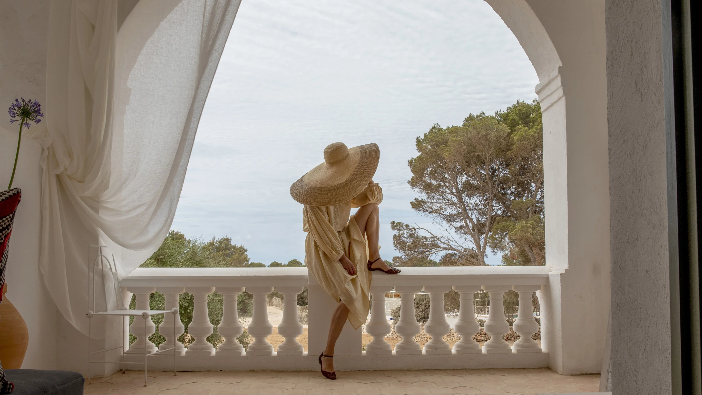 Person in a beige dress and large straw hat sitting on a white balcony railing overlooking trees.