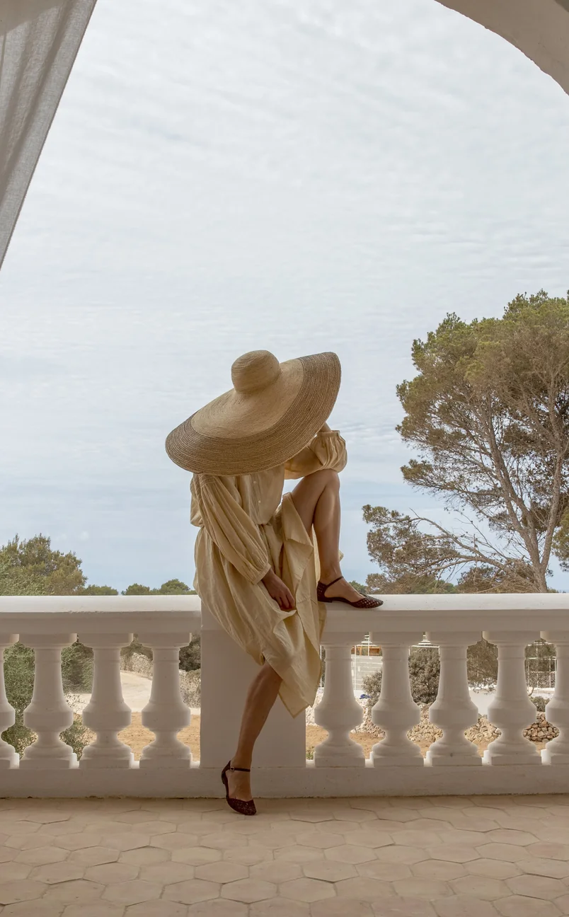 Person in a light beige dress and oversized wide-brimmed straw hat sitting on a white balcony railing with trees in the background.