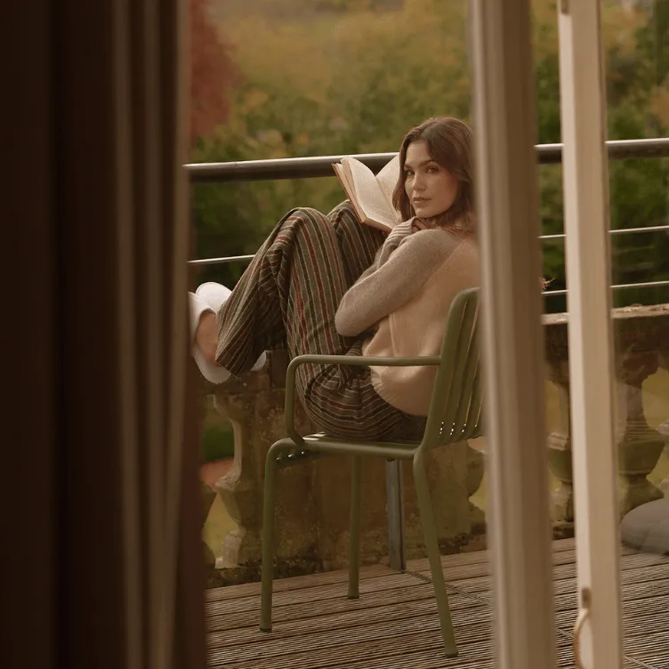 Woman sitting on a green chair on a balcony, reading a book with her legs curled up, surrounded by autumnal trees.