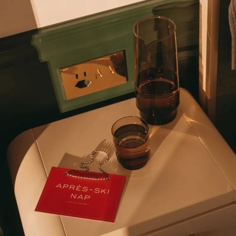 Un vaso y una jarra con líquido ámbar sobre una mesa de color beige junto a una etiqueta roja que dice «APRÈS-SKI NAP».