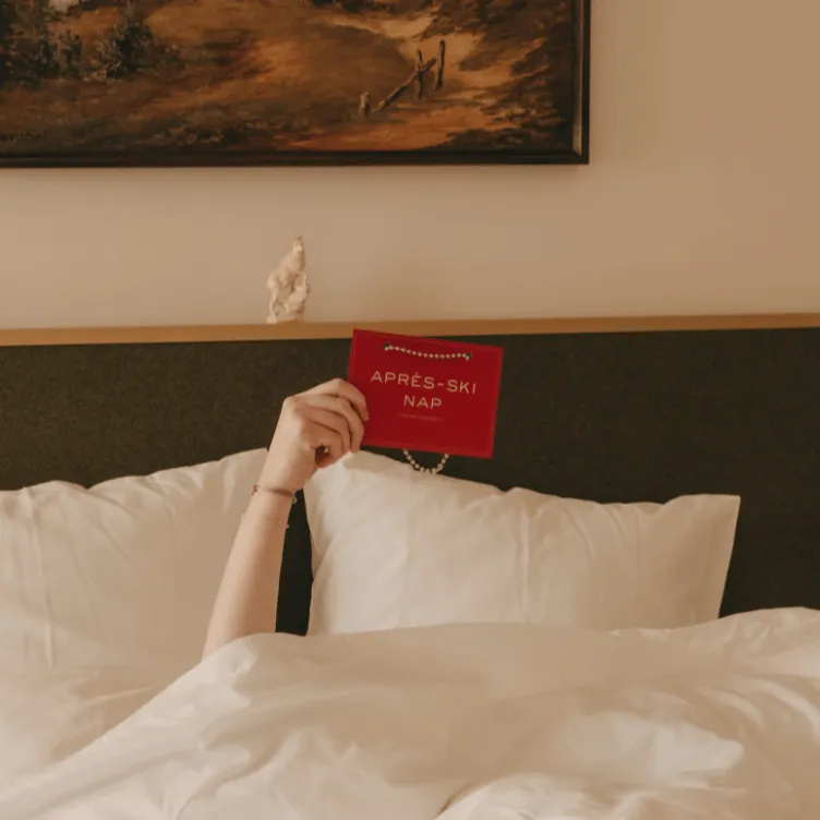 Persona acostada en la cama bajo sábanas blancas con un cartel rojo que dice «SIESTA DESPUÉS DE ESQUIAR».