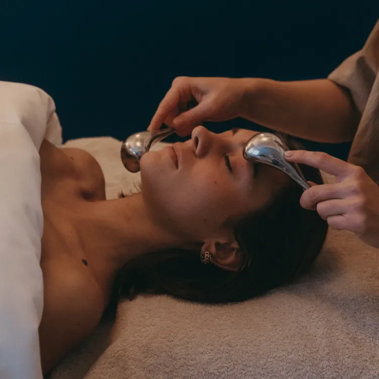 Mujer que recibe un masaje facial con dos herramientas de masaje metálicas en las mejillas mientras está acostada en una cama de spa.