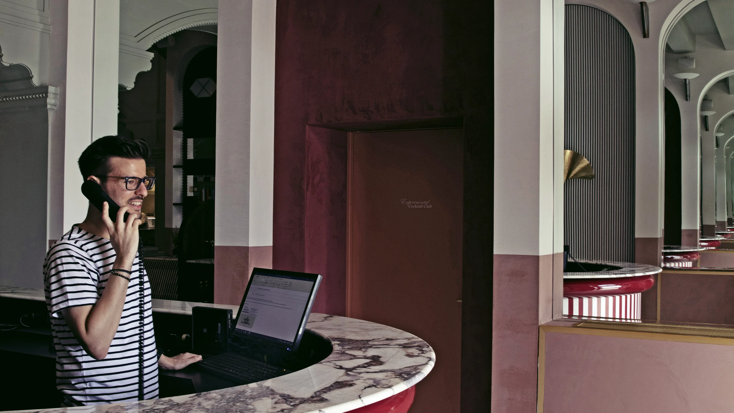 Man in glasses and striped shirt speaking on a corded phone behind a marble reception desk with a computer in a stylish interior.