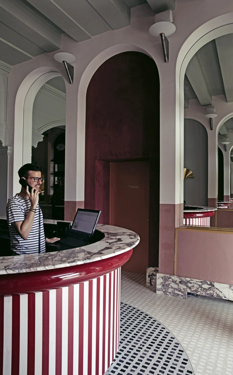 Man wearing glasses and a striped shirt talking on the phone behind a curved reception desk with red and white stripes and marble countertop in a stylish lobby.