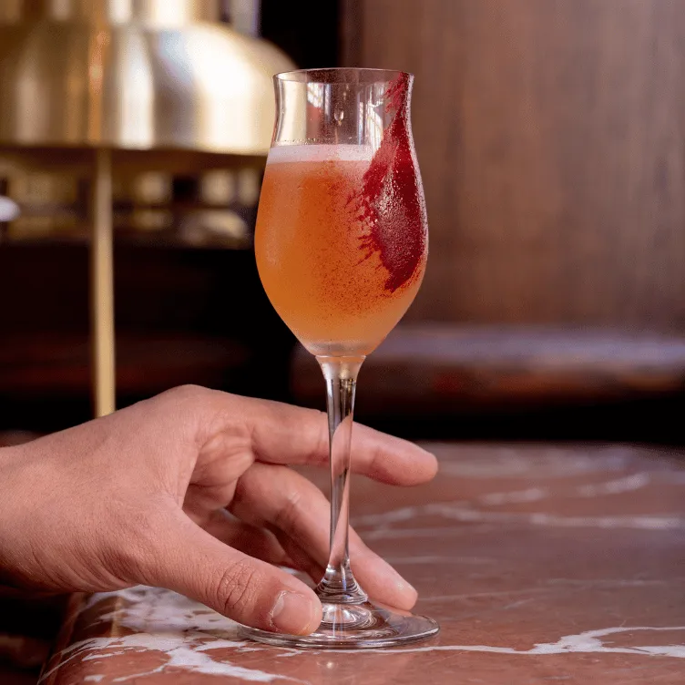 Hand holding a stemmed glass with an orange cocktail garnished with a red fruit or syrup smear.
