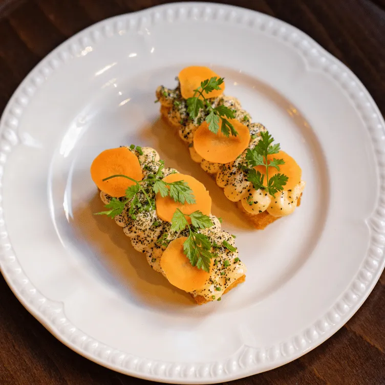 Two rectangular savory appetizers with creamy topping, orange slices, and green herbs on a white plate.