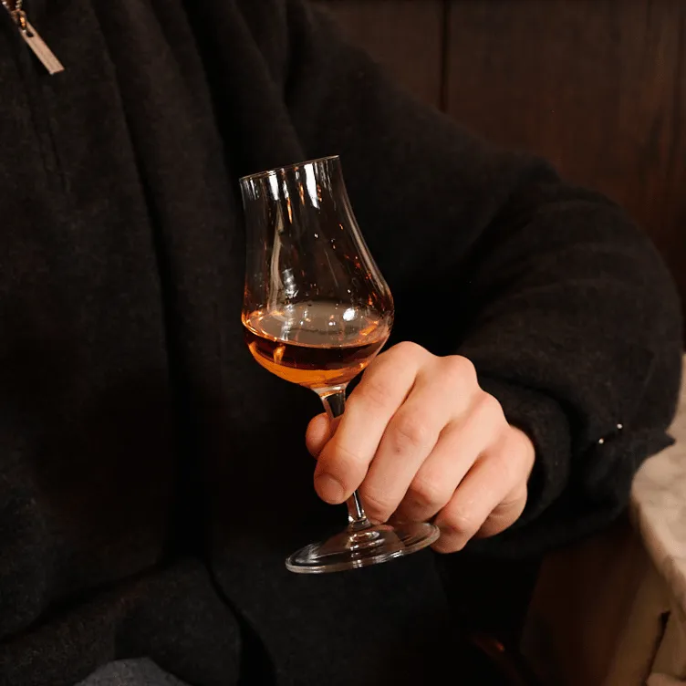 Person wearing a dark coat holding a glass of amber-colored whiskey or brandy.