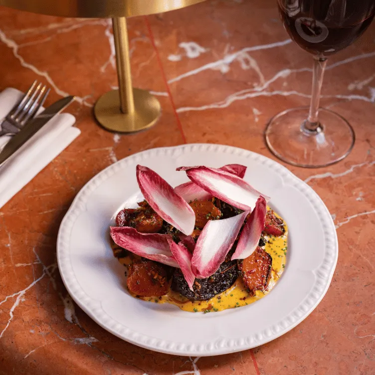 Gourmet dish with grilled vegetables and red chicory leaves on a white plate, served on a marble table with a glass of red wine and cutlery wrapped in a white napkin.