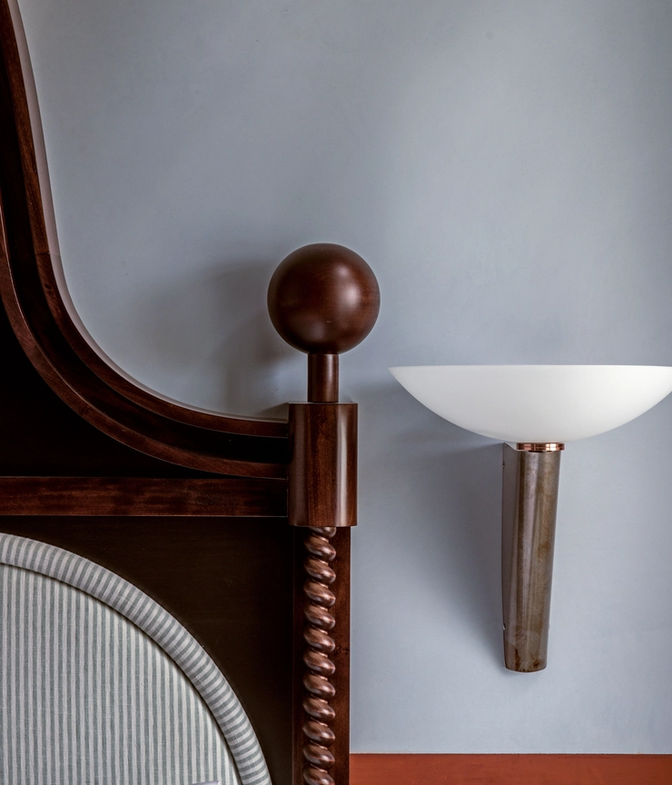 Close-up of a wooden bedpost with a round finial and a striped upholstered headboard beside a modern wall-mounted light with a frosted glass shade.