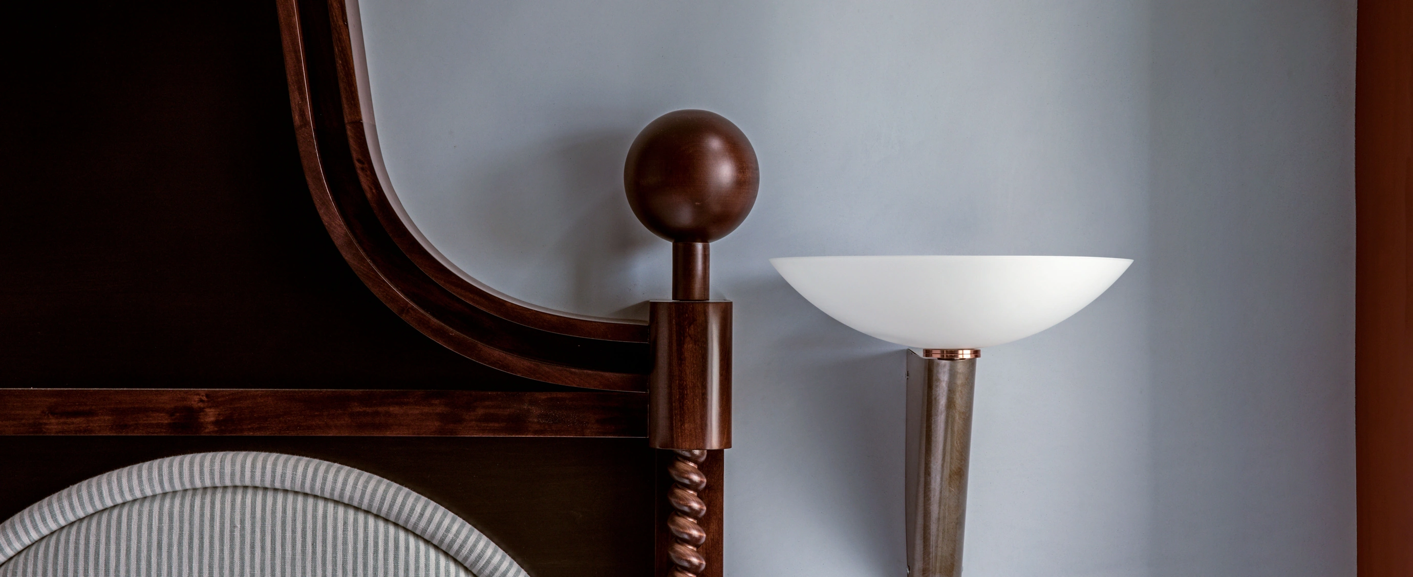 Close-up of a wooden bedpost with a round finial and a striped upholstered headboard next to a modern wall-mounted light fixture with a white bowl-shaped shade.