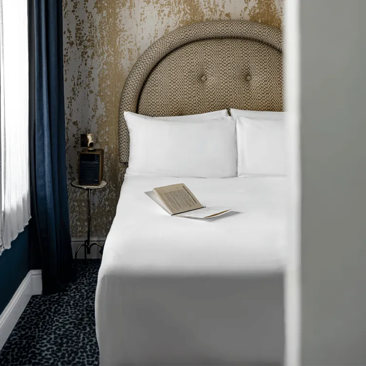 Cama cuidadosamente hecha con sábanas blancas, dos almohadas, un libro abierto en la parte superior, una cabecera redondeada estampada y una mesa auxiliar redonda con altavoz y lámpara junto a una ventana con cortinas transparentes y cortinas oscuras.