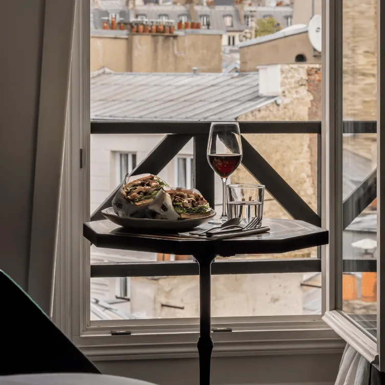 Plato con un sándwich envuelto, una copa de vino tinto y un vaso de agua en una mesa pequeña junto a una ventana abierta con vista a los tejados.