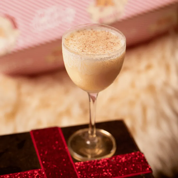 Creamy cocktail in an etched glass with a sprinkle of nutmeg on top, placed on a dark surface with a red glittery ribbon.