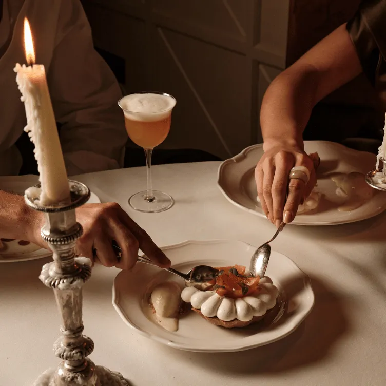 Two people sharing a dessert with a whipped cream topping and fruit on a white plate at a candlelit table with a cocktail glass.