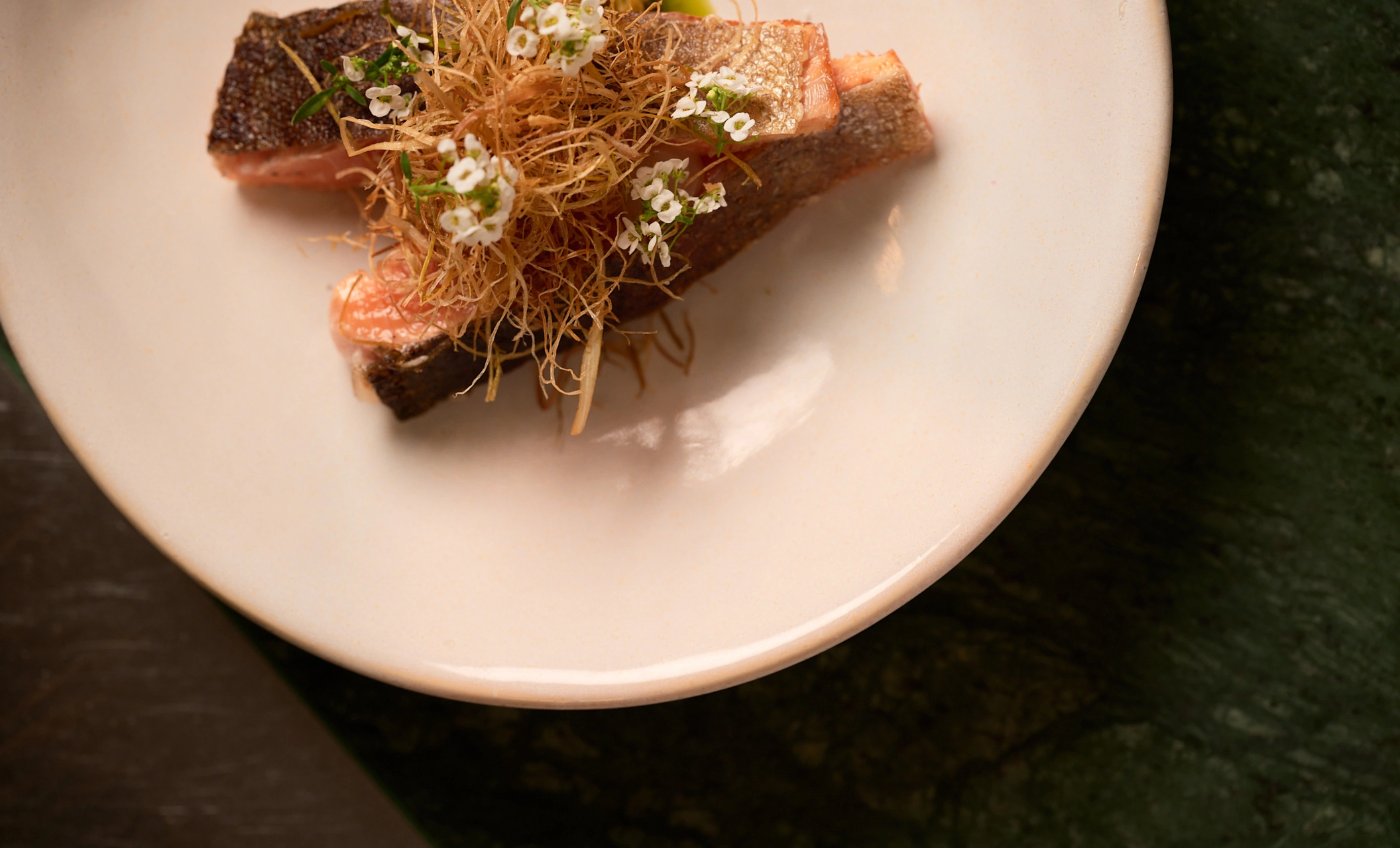 Seared salmon fillet topped with crispy fried garnish and small white edible flowers on a white plate.