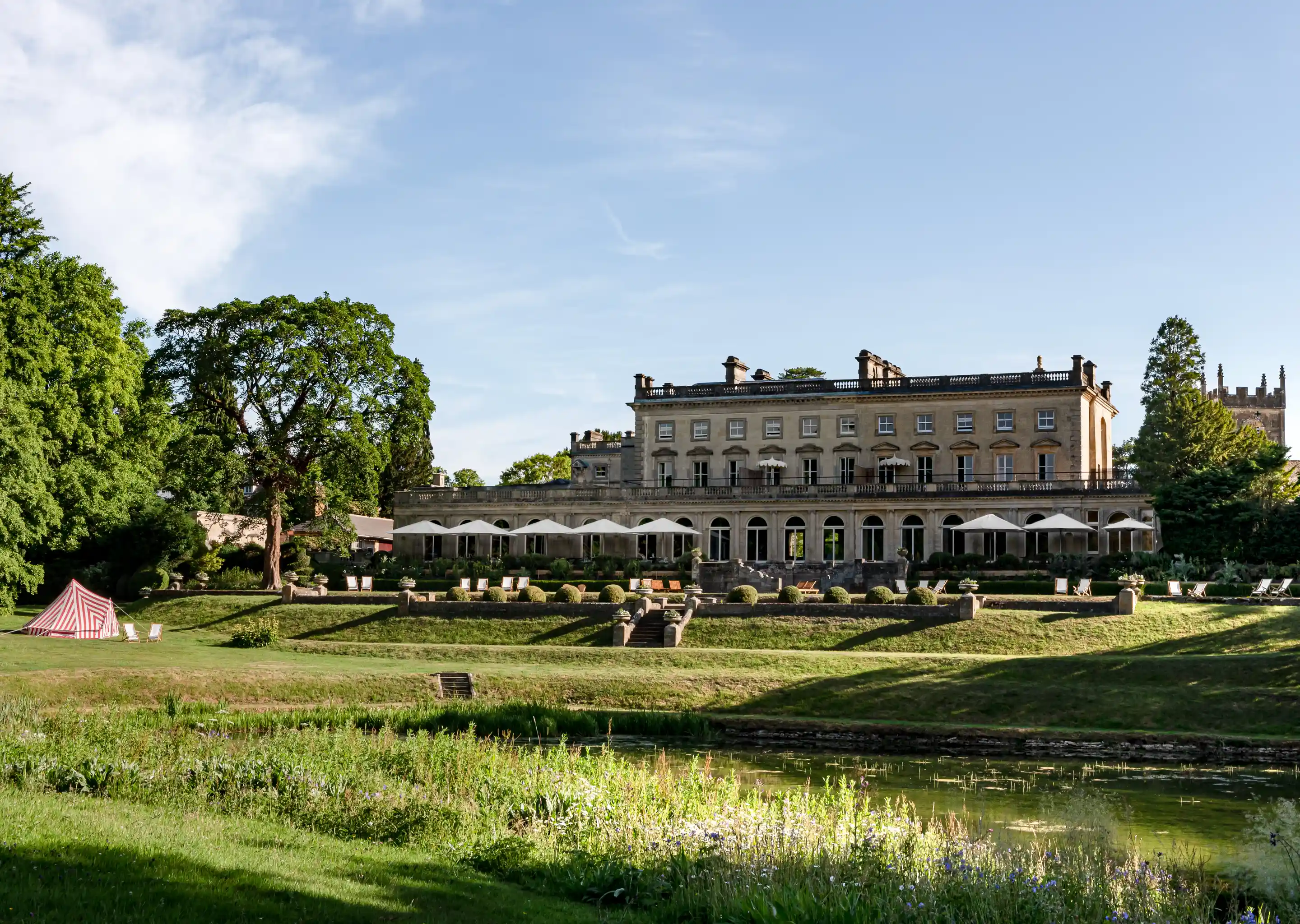 Large historic stone hotel building with white umbrellas on the terrace, surrounded by green lawns, trees, and a pond in the foreground under a clear sky.