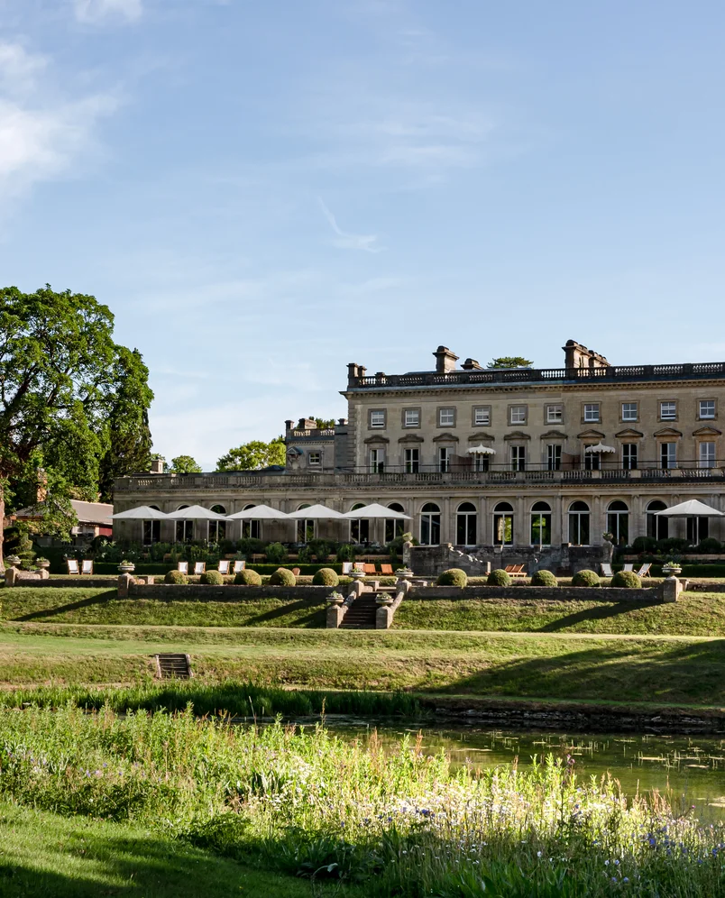 Elegant historic hotel building with large windows, outdoor seating areas with umbrellas, manicured gardens, trees, and a pond in the foreground under a clear blue sky.