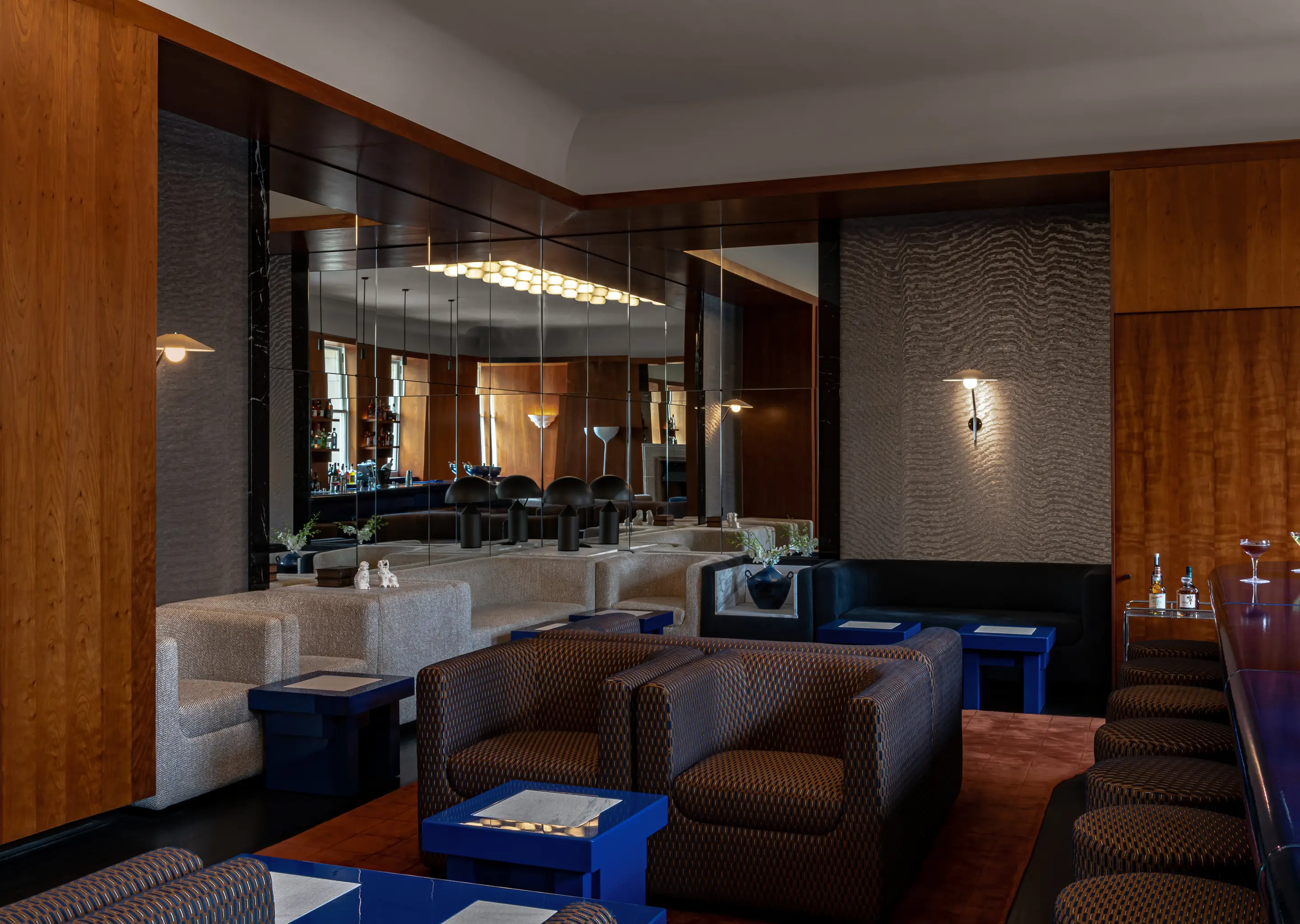 Sophisticated cocktail bar lounge with patterned armchairs, blue side tables, wood-paneled walls, and a large mirror reflecting the space.