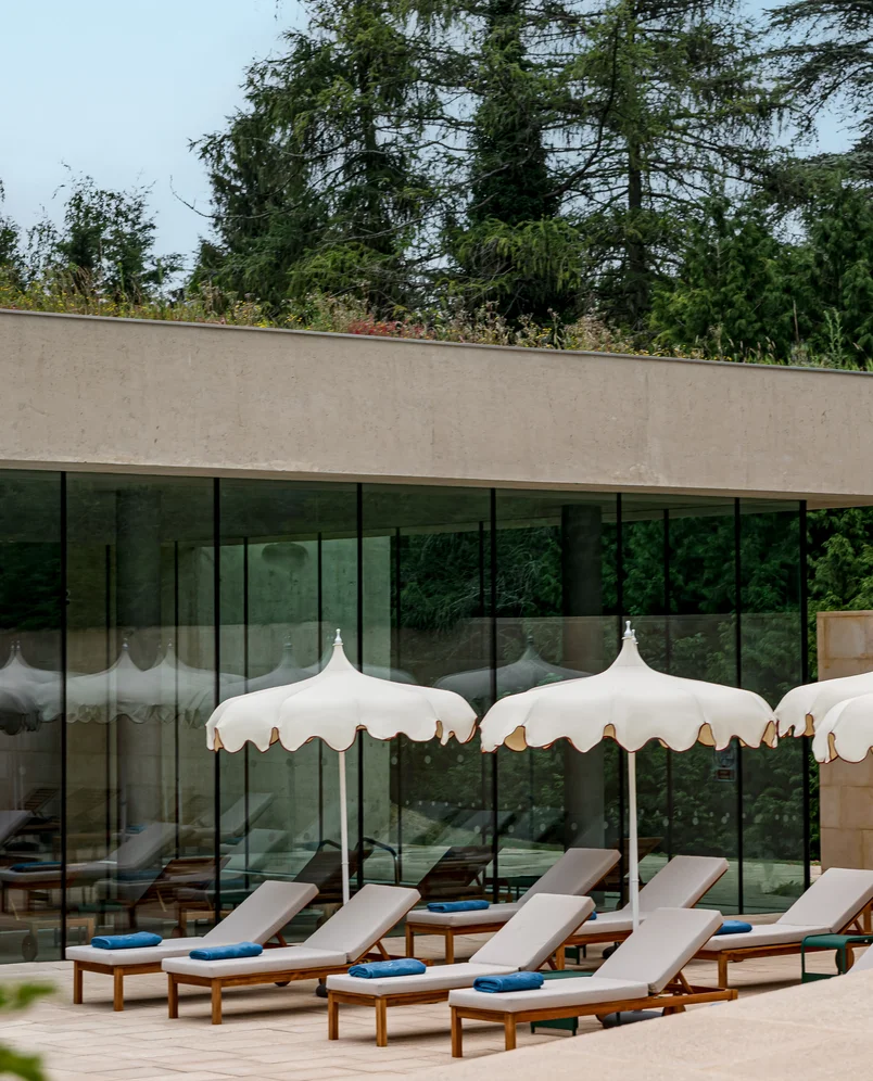 Outdoor lounge chairs with blue towels under white scalloped umbrellas in front of a building with large glass windows surrounded by trees.