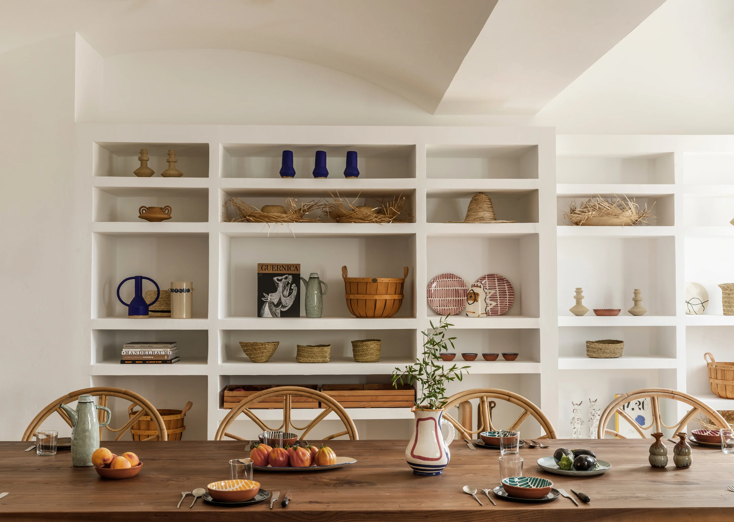 Dining table set with assorted fruit and pottery in front of white built-in shelves holding decorative items including baskets, plates, and vases.