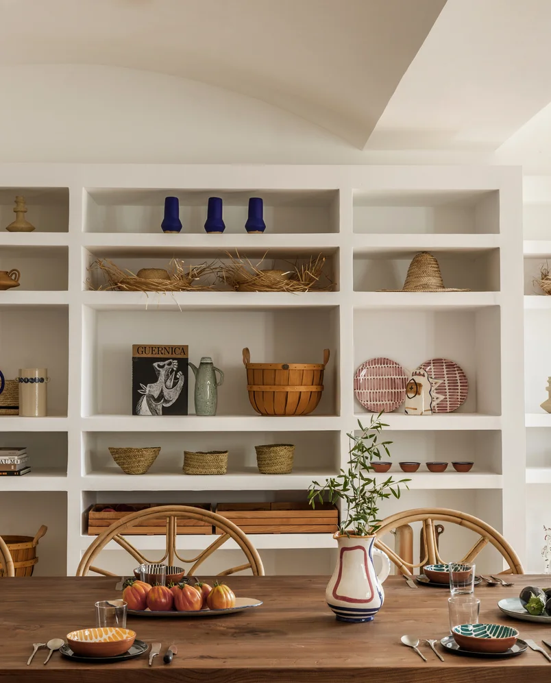 Dining area with wooden table, two rattan chairs, ceramic plates, glasses, and a vase with greenery in front of built-in white shelves holding decorative baskets, pottery, hats, and a book titled 'Guernica'.
