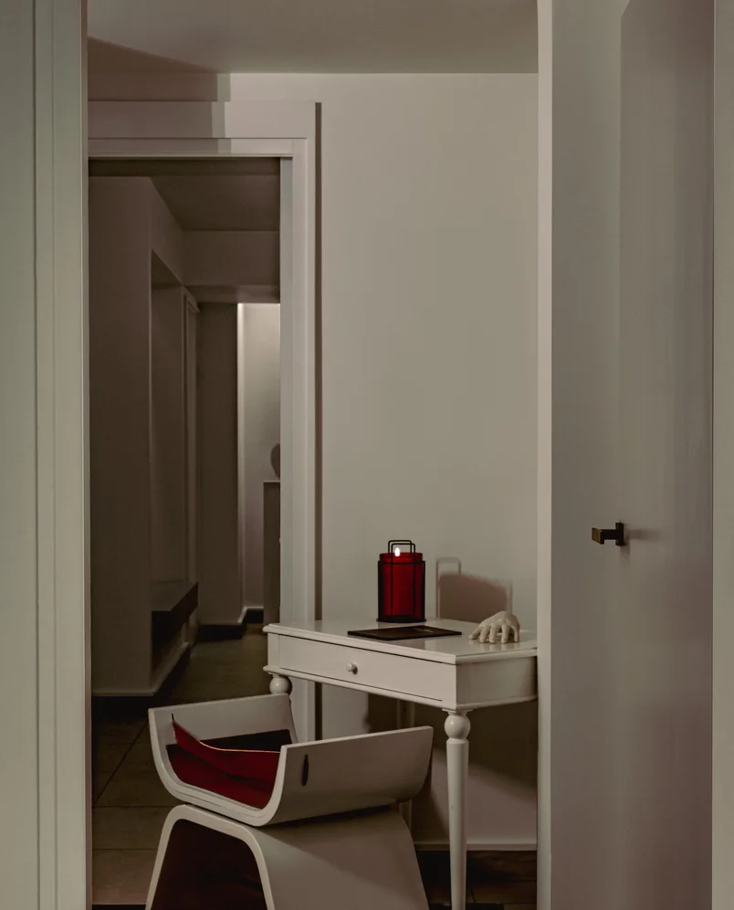 White desk with a red lantern and a decorative white hand sculpture, accompanied by a modern white chair with a red cushion in a minimalistic hallway.