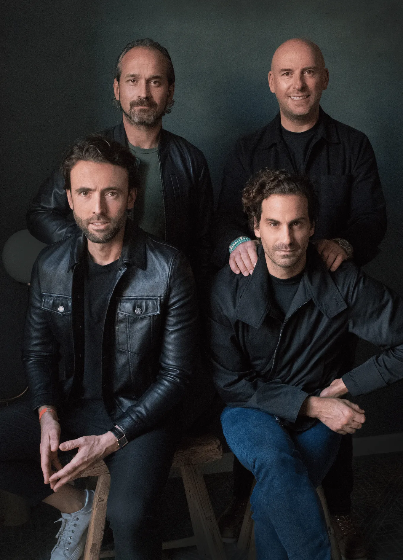 Group portrait of four men wearing dark jackets sitting and standing against a dark background.