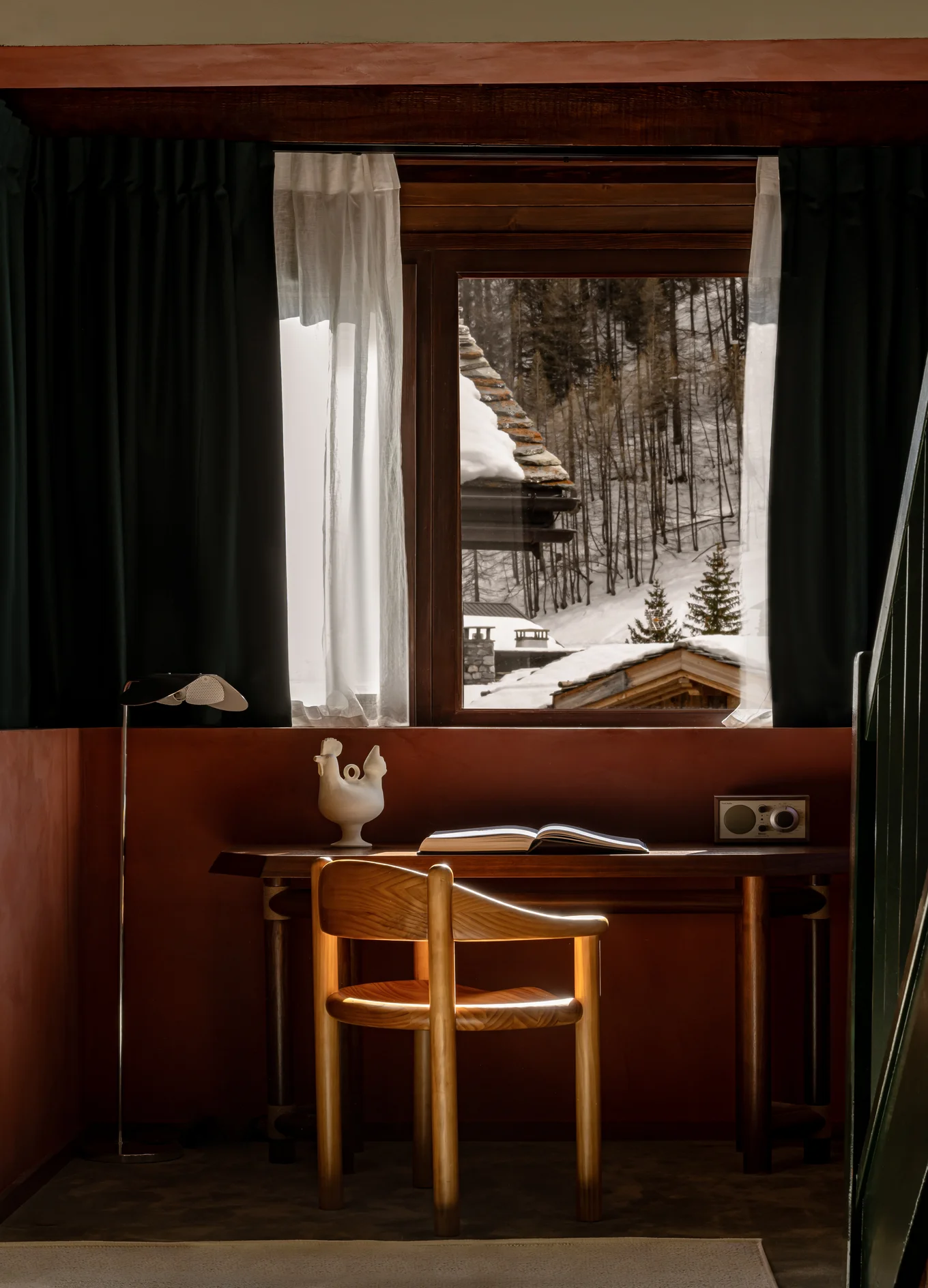 Cozy wooden desk and chair by a window with translucent curtains, overlooking snowy rooftops and forest.
