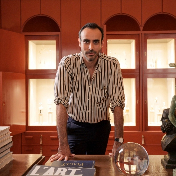 Man with striped shirt leaning on a wooden table with books and a glass sphere, standing in front of a red cabinet with illuminated display cases.