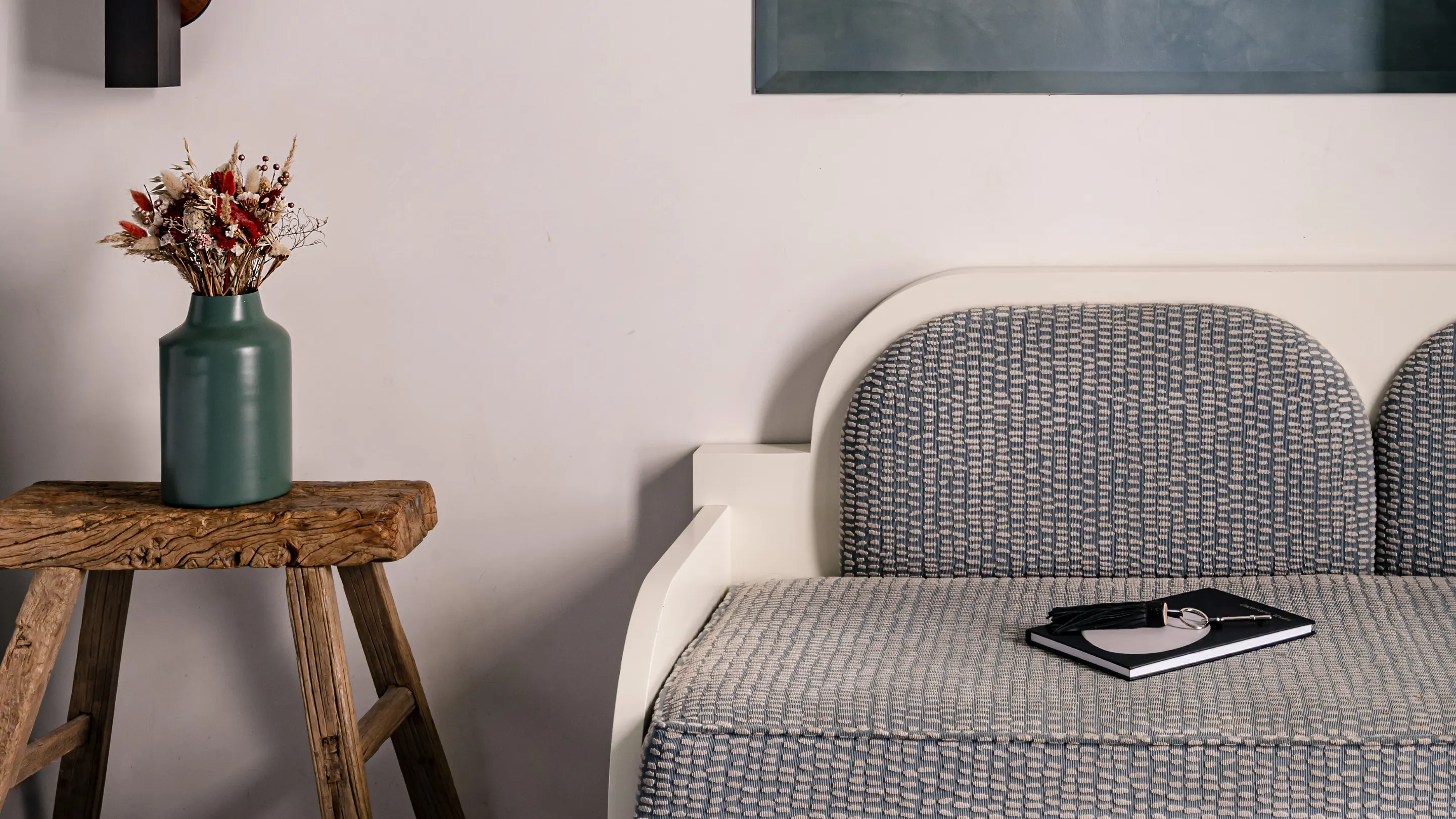 Rustic wooden stool with green vase holding dried flowers beside a white and gray patterned sofa with a notebook and tassel on it.