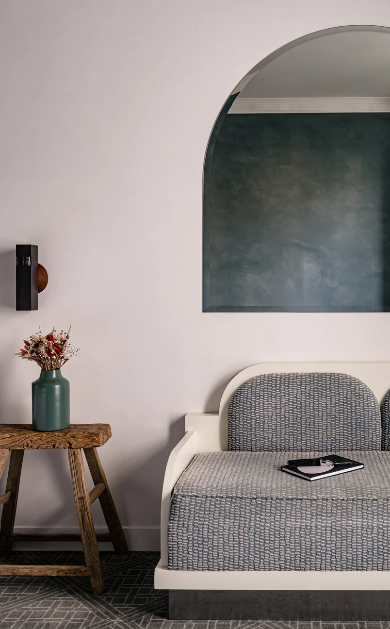Minimalist room corner with a wooden bench holding a green vase of dried flowers, next to a white-framed patterned sofa with a notebook and glasses placed on it, and a large arched mirror on the wall.