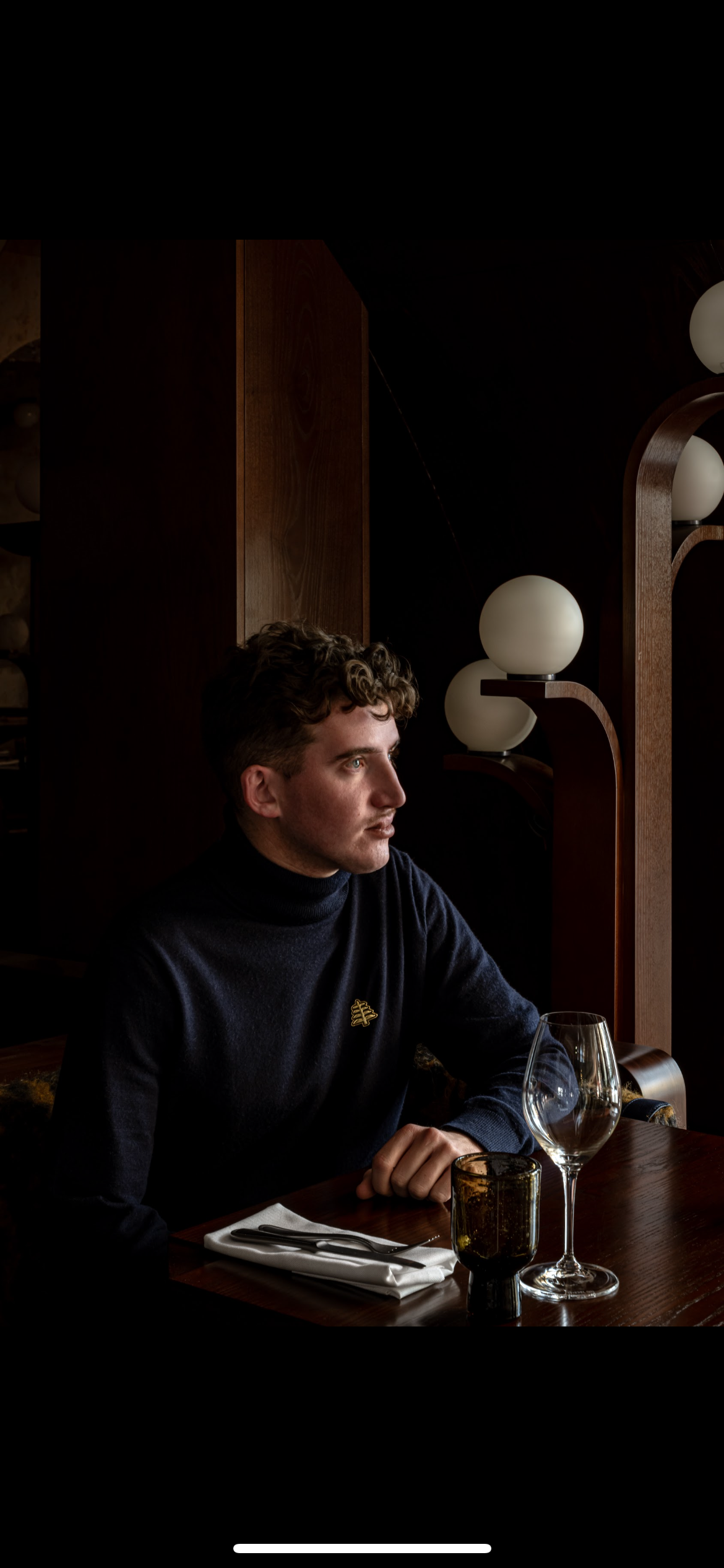 Young man with curly hair wearing a dark turtleneck sitting at a wooden table with a glass and cutlery in a dimly lit restaurant.