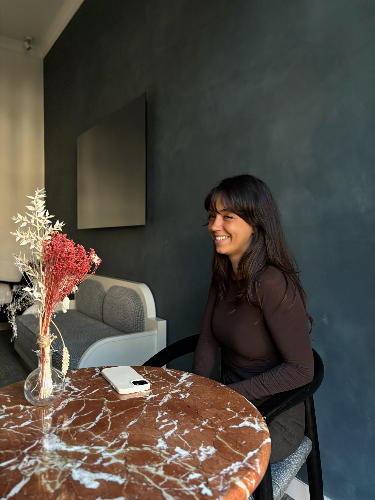 Woman with long dark hair sitting at a marble table with a vase of dried flowers and a smartphone, smiling against a dark blue wall.