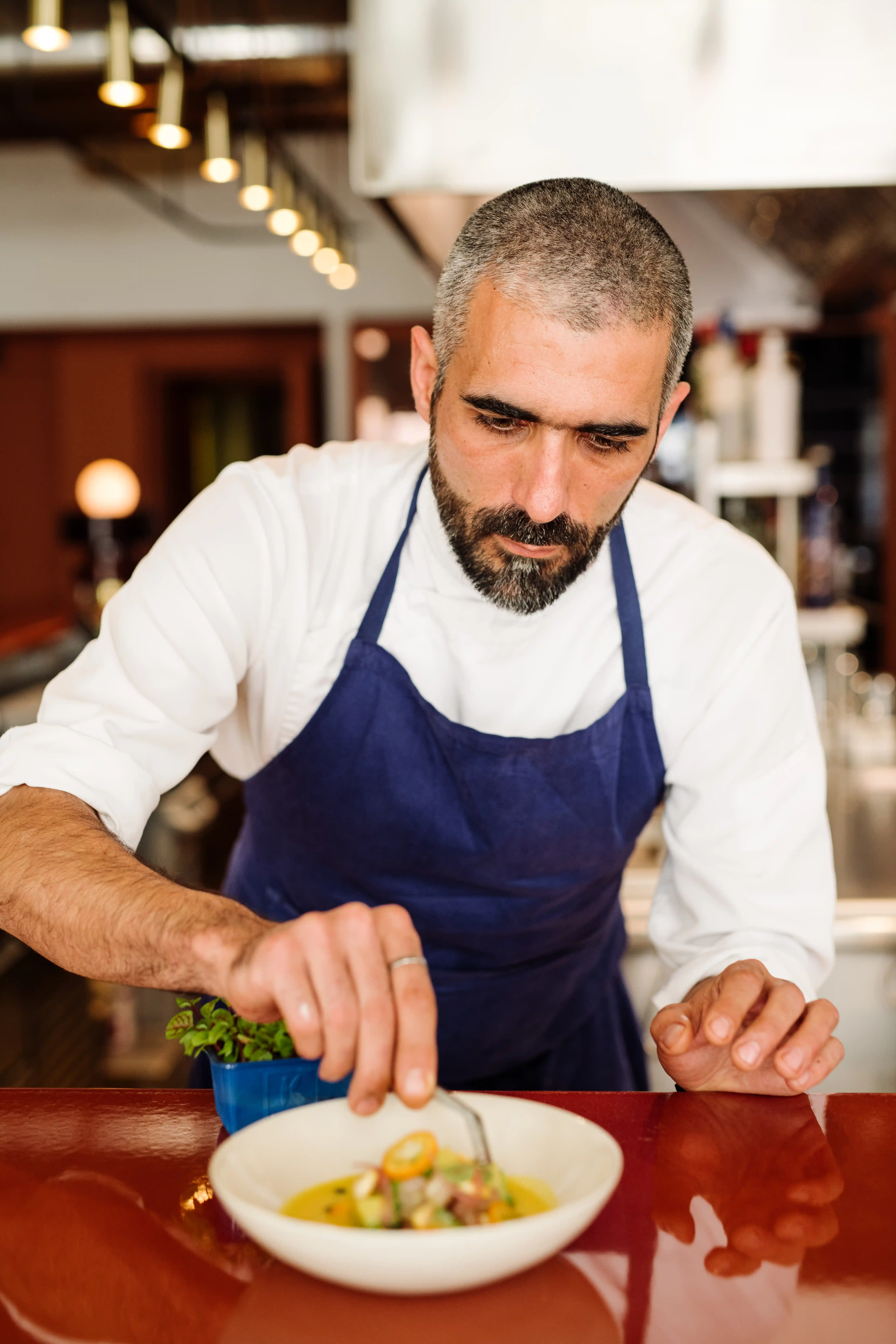 Male chef in a blue apron carefully plating a dish in a restaurant kitchen.