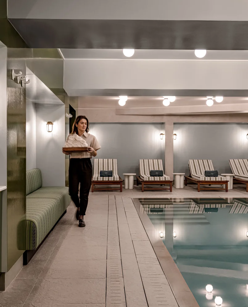 Woman carrying folded towels walking beside an indoor pool with lounge chairs and soft lighting.